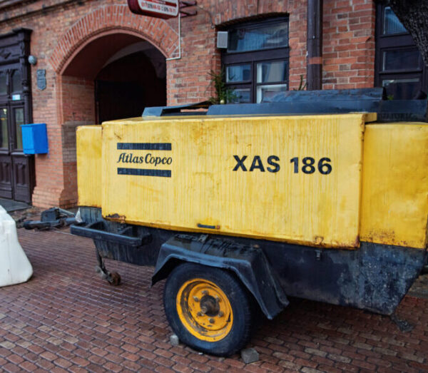 Minsk, Belarus. Dec 17, 2024. Yellow Atlas Copco XAS 186 air compressor on trailer parked on brick paved sidewalk beside old red brick building. Air compressor, construction and maintenance tasks.
